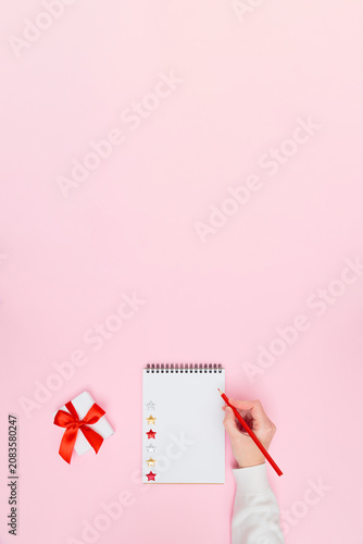 Female hand writing on blank notepad with gift box and red ribbon on pastel pink background, festive flat lay vertical banner with copy space for holiday promotion, planning, goals, to-do list.