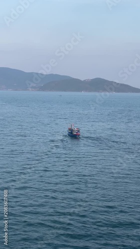 Small fishing boat crossing calm teal sea with clear wake, distant low hills, pale sky and textured water, creating peaceful maritime scene with strong diagonal movement and open horizon