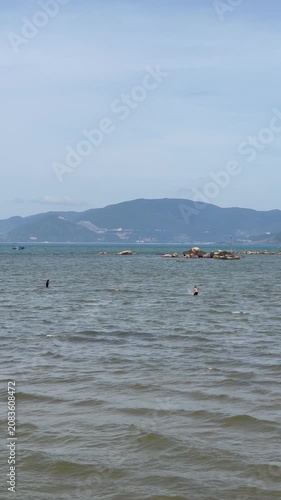 Two people moving through shallow coastal water beside rocky islet, with distant mountain range, calm horizon, small moored boat, pale blue sky, and layered seascape in soft daylight