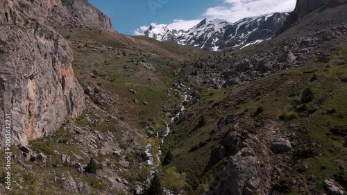 Rocky cliffs rise on either side of a narrow valley. Clear stream flows through green vegetation. Sunlight glints off the water, highlighting rugged terrain