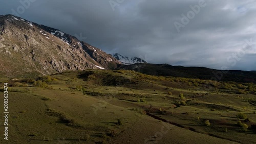 Snow-capped mountains rise behind a grassy plain. Dark storm clouds cast dramatic shadows. The landscape is empty of people or animals. Ideal for nature documentaries or travel advertising