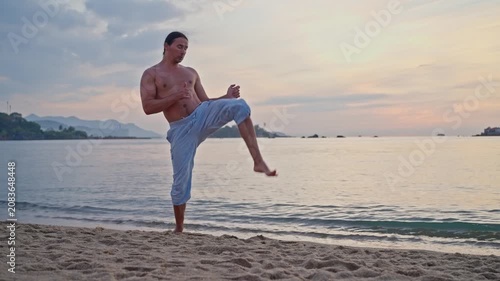 Shirtless man practicing martial arts on sandy beach at sunset, moving through balanced kicks and upright recovery beside calm sea, pastel sky, warm light, distant islets