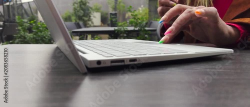 Adult Female Hand with Colorful Manicure and Multicolored Nail Polish Using Laptop Trackpad on Portable Computer at Modern Outdoor Workspace on Wooden Table with Visible Cigarette Smoke 
