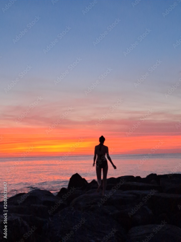 Fototapeta premium Silhouette of a woman standing on the rocks watching an incredible vibrant orange sunset over the sea.