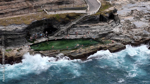 Aerial view of Bogey Hole ocean pool carved into rock with people swimming in clear green water, surrounded by rugged cliffs and crashing waves, Newcastle, New South Wales, Australia.