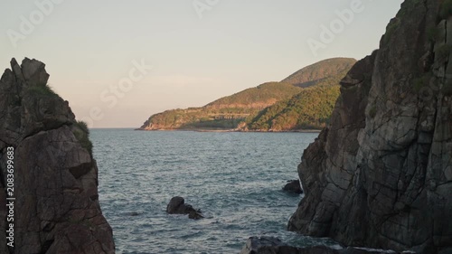 Nha Trang Vietnam. Panoramic coastal inlet framed by jagged cliffs with choppy blue-gray sea and illuminated tree-covered headland under warm