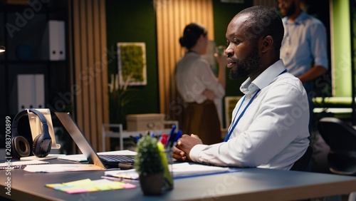 фотографія Black man accountant in office using laptop and writing down information, working late to finish tasks before deadline, transcribing from screen and taking notes, documenting research