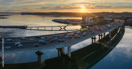 Fotografie Auckland, New Zealand - Commuters drive on the Mangere Bridge during sunset, with the city skyline and harbor in the background