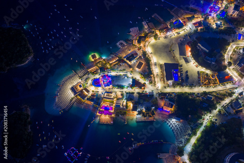 Elevated night drone shot of coastal road, parked cars, glowing resorts, and illuminated beach clubs extending into the sea at Ksamil, Albanian Riviera. Mediterranean holiday.