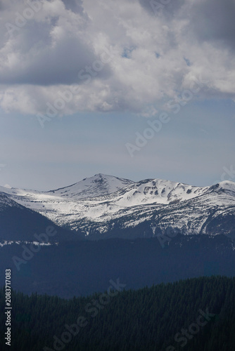 Vertical scenic landscape of snow covered mountain ridge under dramatic cloudy sky with forest below.