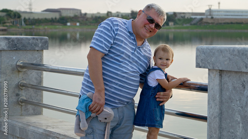 Obraz na plátně Happy mature father or grandfather hugging his cute toddler daughter or granddaughter outdoors by the lake
