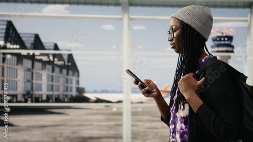 Woman holds phone inside modern airport terminal, doing online check-in, while girl uses smartphone to access internet airline website and check plane timetable.