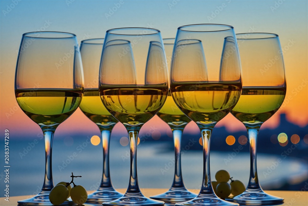 Obraz premium Six glasses filled with white wine stand against dusk ocean sunset sky. Grapes and city lights form background. Perfect for celebrations or quiet relaxation.