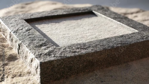 A close up view of a square stone frame filled with fine sand, resting on a textured rock surface. Natural sunlight highlights the rough and smooth textures in this minimalistic composition