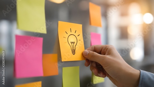 Agile planning workshop showing a hand attaching a yellow lightbulb sticky note to a glass wall during a brainstorming session.