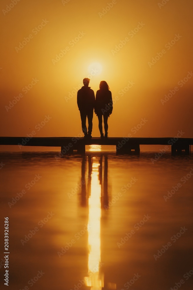 Fototapeta premium Couple stands on a pier at sunset with the sun glowing in the background while the water reflects light