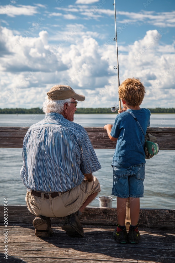 Fototapeta premium Man teaches young boy how to fish from pier during a sunny day with clouds in the sky by a calm body of water