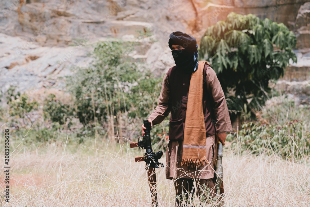 Fototapeta premium Outdoor military concept. Masked armed man in dry grass near rocky cliff, rural conflict scene