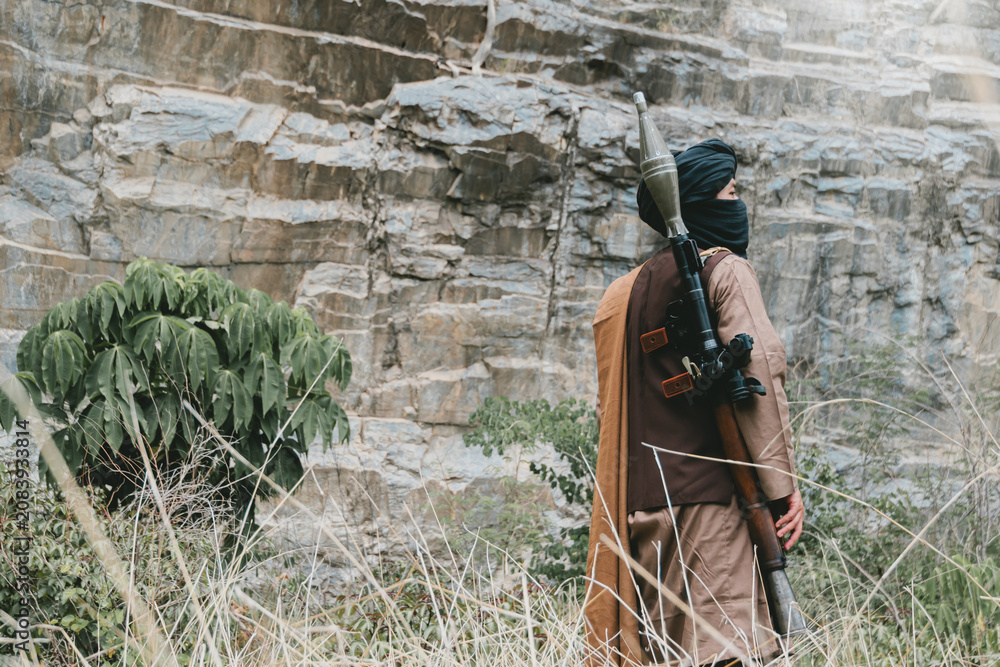 Fototapeta premium Outdoor military concept. Desert warrior with rocket launcher stands near rocky cliff in dry grass