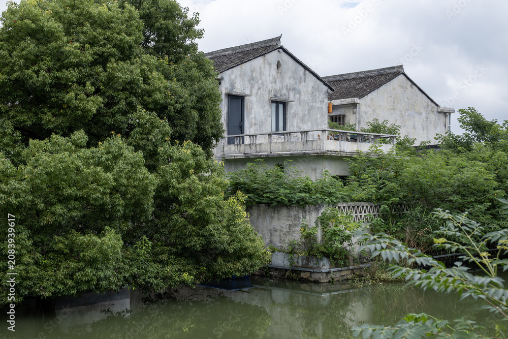 Fototapeta premium Weathered Jiangnan whitewashed house beside a calm river surrounded by lush vegetation in rural Zhejiang China. Tranquil riverside dwelling, cultural heritage, rural homes, traditional lifestyle