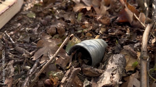 Metal fragment of a cluster munition lying in oak forest leaf litter, with marker stakes placed nearby during humanitarian demining — a poignant editorial visual of postconflict hazards