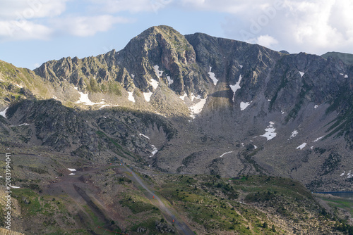 A dramatic Pyrenean landscape of rocky summits and snow-filled gullies rises above a switchback mountain road in Andorra. Sparse alpine vegetation dots the rugged terrain beneath a bright summer sky