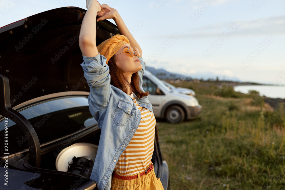 Fototapeta premium Cheerful woman outdoors, stretching with arms raised near an open trunk, sunlit countryside, striped top, denim jacket, carefree mood, natural light, relaxed lifestyle and breeze