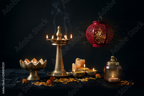 A serene display of glowing traditional brass oil lamps and diyas alongside a vibrant red decorative lantern with illuminated candles and scattered orange marigold petals