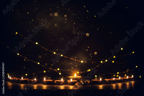 A row of radiant traditional clay oil lamps burn brightly with warm flickering flames illuminating a dark space adorned with sparkling golden bokeh lights and scattered glowing particles