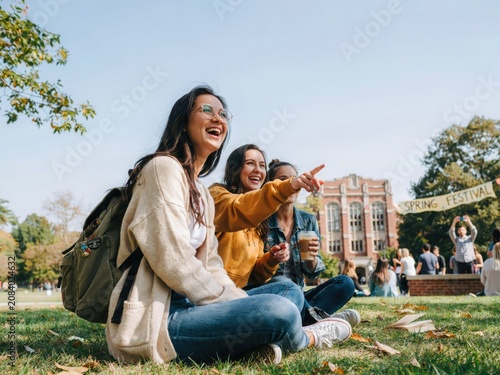 Friends laughing and pointing on university campus lawn during sunny day.