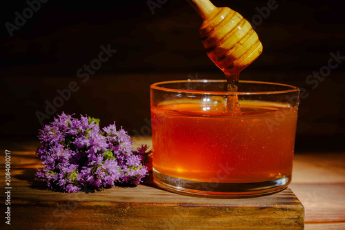 Honey dipper pouring golden amber honey into a glass jar, next to a bunch of fresh purple thyme wildflowers on a wooden surface