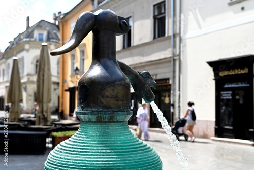 soda syphon bottle sculpture and fountain in public square. streetscape in Gyor, Hungary. layered green glass in shallow blue pool. popular landmark. outdoors, travel, tourism concept. people walking 