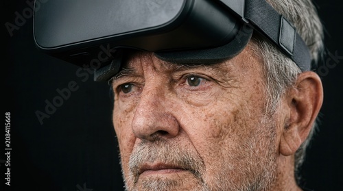 Elderly man wearing virtual reality headset with gray hair and facial wrinkles, focused expression, against a dark background highlighting technology and innovation