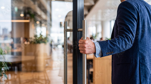 Photos Close-up of a businessman's hand opening a sleek glass door to a modern, brightly lit corporate office