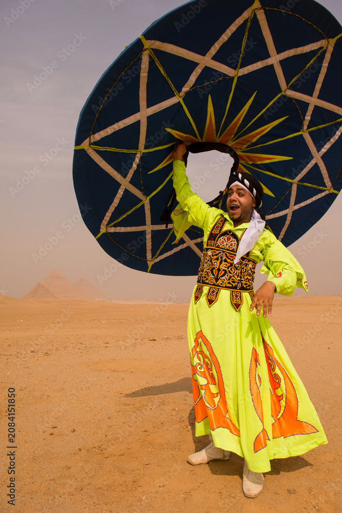 Fototapeta premium Tanoura dancer in bright neon embroidered robe raises spinning disc overhead with joyful expression before Great Pyramids of Giza ideal for cultural