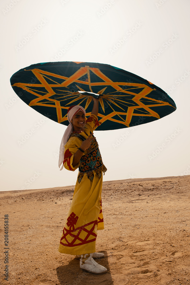 Fototapeta premium Nubian folk performer wearing vibrant embroidered yellow robe and red turban smiles while raising large geometric teal star pattern umbrella in open