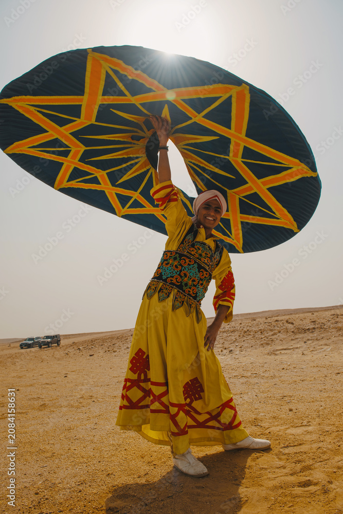 Fototapeta premium Joyful folk dancer in embroidered yellow traditional costume raises a bold teal and orange star patterned ceremonial umbrella overhead in a vast sunny