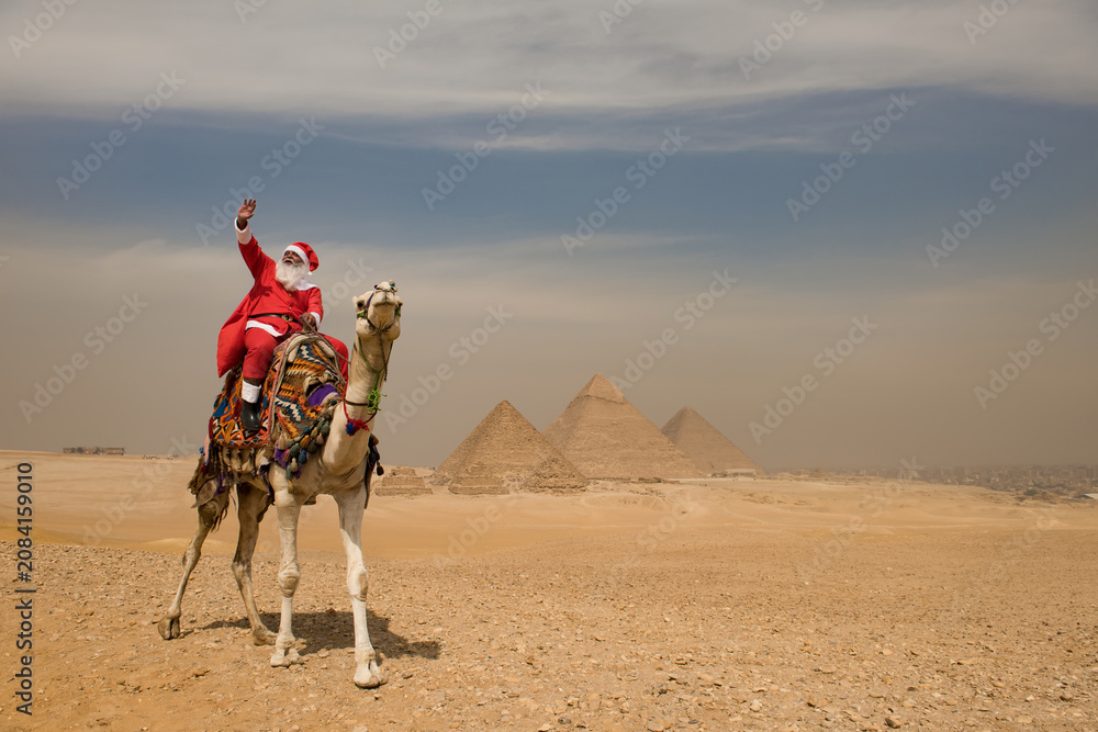 Fototapeta premium Joyful Santa Claus riding a colorfully decorated camel and waving high before the three great Giza pyramids across a sunlit Egyptian desert plateau