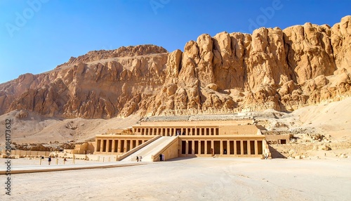 A large, ancient structure with pillars and ramps, set against a towering rock formation under a clear blue sky. It is located in a desert