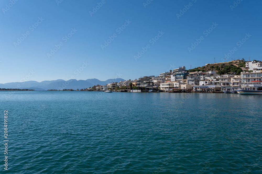 Fototapeta premium Waterfront buildings of Elounda coastal town line Mirabello Bay shoreline on Crete island under sunny blue sky with mountains.
