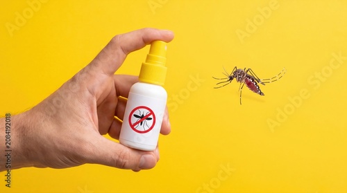 Hand holding mosquito repellent spray bottle against a flying insect on yellow