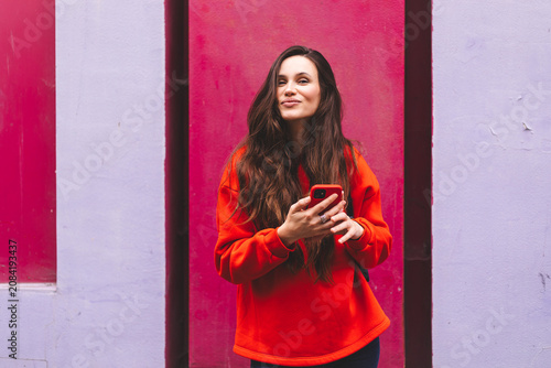 Playful young woman in red sweatshirt using mobile phone against colorful purple and pink wall background, beautiful brunette girl with funny facial expression holding smartphone city street portrait