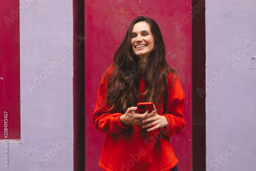 Happy young woman laughing at camera while holding smartphone against colorful purple and pink wall background, beautiful brunette girl in red sweatshirt urban lifestyle technology portrait