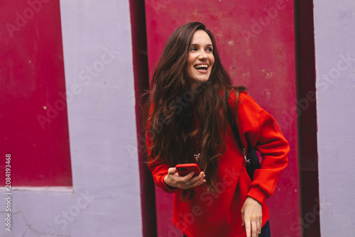 Excited young woman looking away and laughing while holding mobile phone against colorful wall, happy brunette girl in red sweatshirt using smartphone urban lifestyle technology side portrait