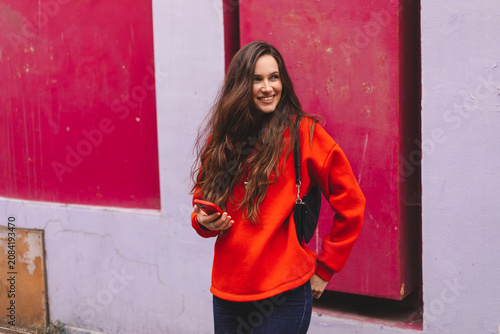 Smiling young woman looking at camera while holding mobile phone against colorful wall background, attractive brunette girl in red sweatshirt using smartphone urban lifestyle technology portrait