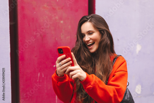 Excited laughing young woman looking at smartphone screen holding mobile phone, happy emotional female influencer reading good news or watching funny content online against magenta wall outdoors