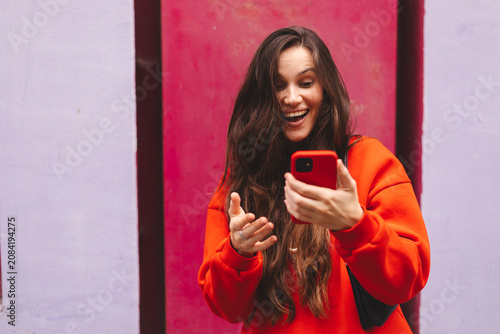 Surprised young woman looking at smartphone screen with open mouth gesturing hand, amazed emotional female influencer reading shocking good news holding mobile phone outdoors against magenta wall