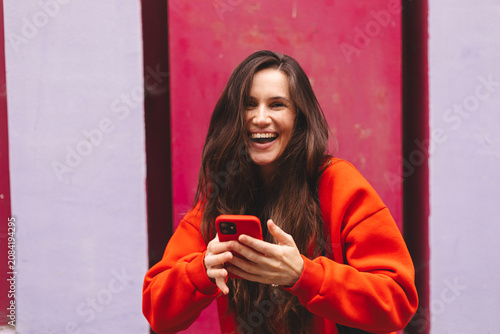 Joyful laughing young woman looking at camera holding smartphone, happy emotional female influencer in red sweatshirt with mobile phone celebrating success outdoors against bright magenta wall