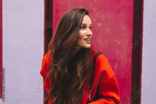 Attractive young woman turning around smiling and looking away, happy half profile view female influencer with long wavy hair posing outdoors against bright magenta wall, candid urban portrait