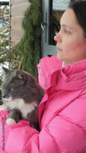 Woman in pink jacket holding gray cat near doorway, looking down at pet while standing outdoors, cozy winter portrait with close view of face, fur, and hands, soft natural light visible outside.
